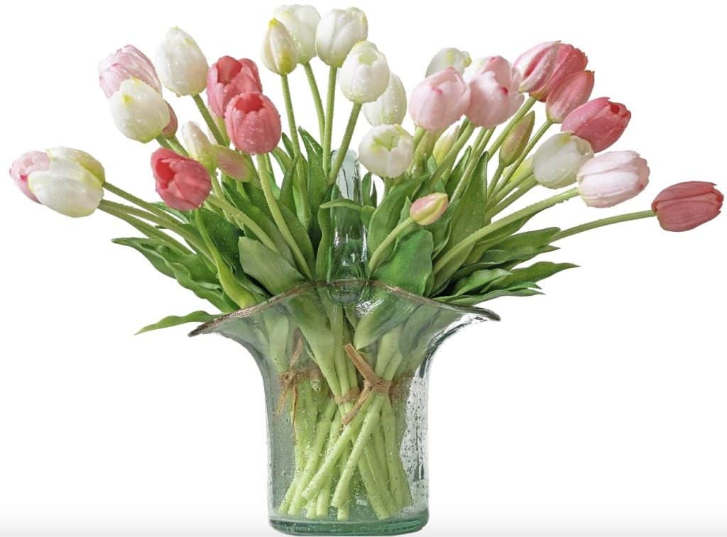 A clear glass vase showcases a large bouquet of pink and white tulips with green leaves, resembling exquisite pink crafts. The vase, with its wide, flared top, is elegantly set against a plain white background.